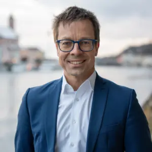 Lächelnder Mann mit blauer Brille und Sakko vor unscharfer Passauer Flusskulisse