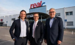 Drei Männer in Anzügen stehen lächelnd vor dem real-Einkaufsmarkt in Passau