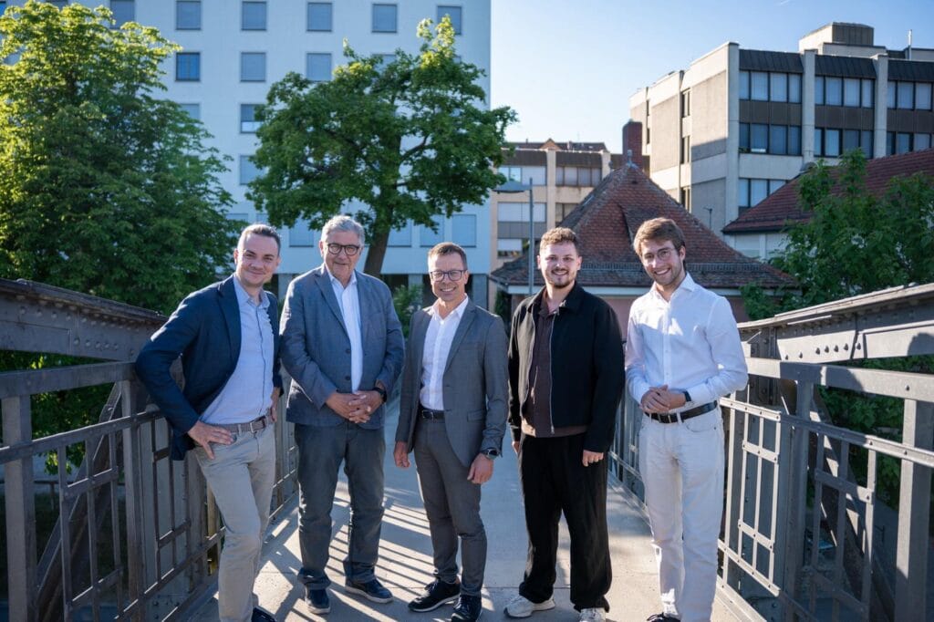 Fünf Männer posieren gemeinsam auf einer Brücke im Freien, im Hintergrund Stadtgebäude und Bäume