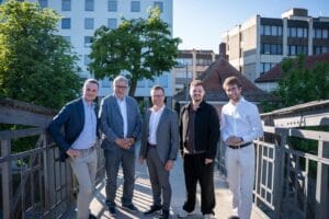Fünf Männer posieren gemeinsam auf einer Brücke im Freien, im Hintergrund Stadtgebäude und Bäume