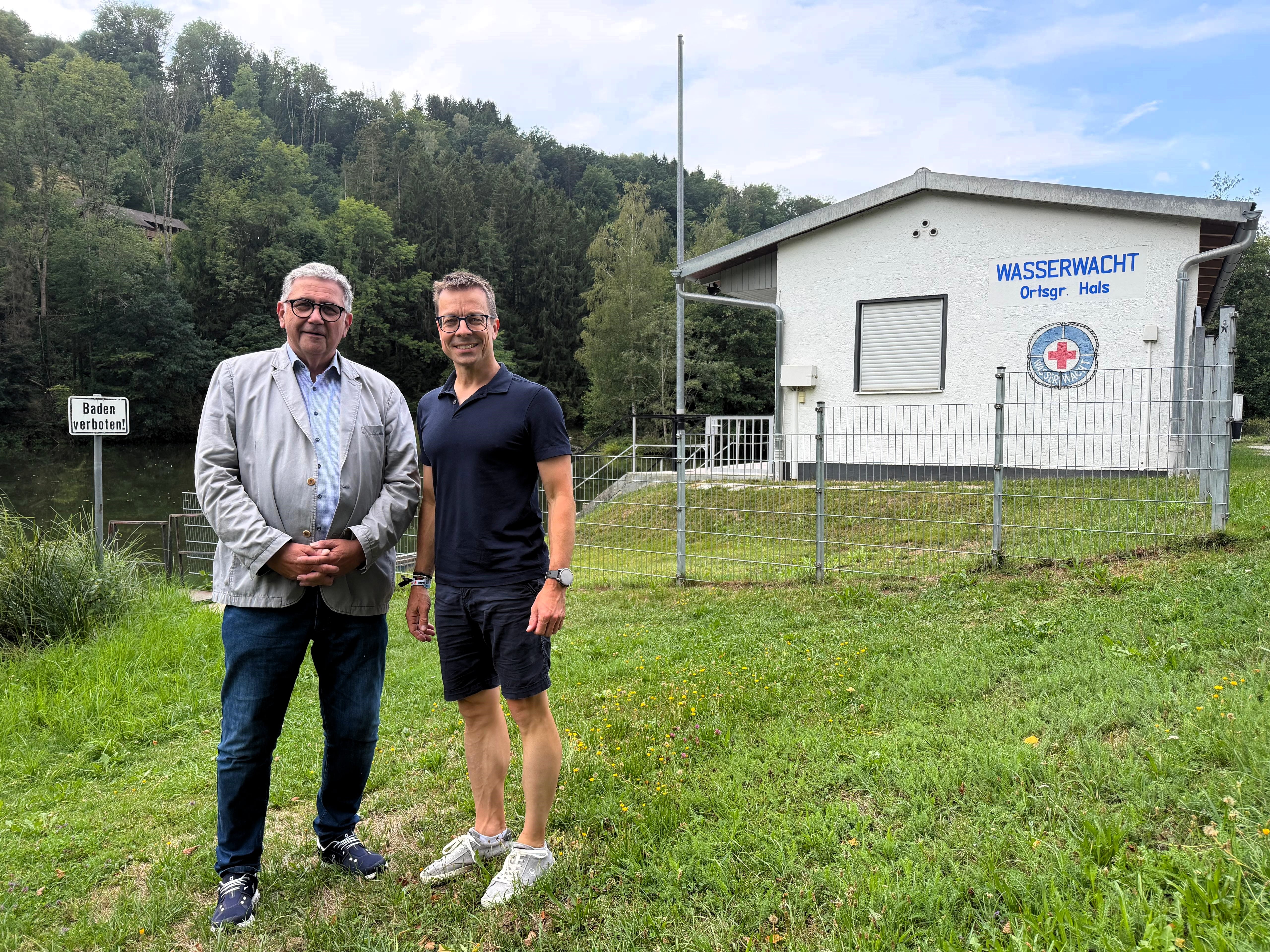 Zwei Stadträte am Halser Stausee neben dem Schild Baden verboten, im Hintergrund das Gebäude der Wasserwacht Ortsgruppe Hals